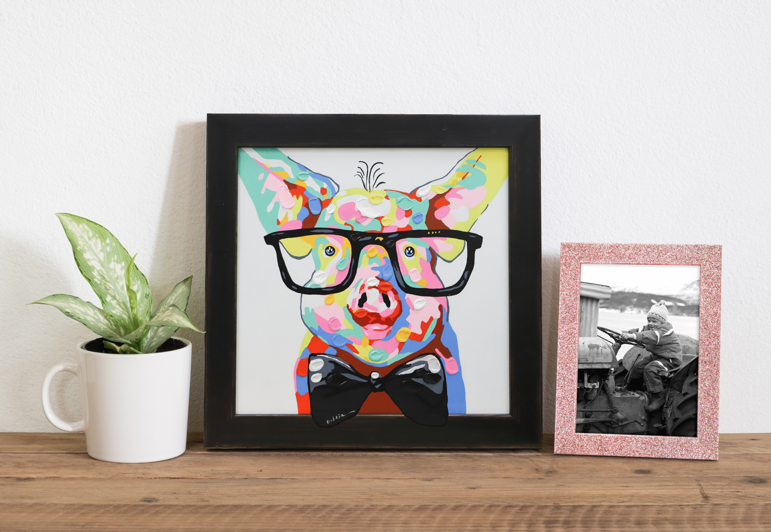 A photo of a painting of a pig framed in a black rustic paired with a pink sparkly frame featuring a photograph of a little girl. Both frames are displayed on a shelf with a small plant.