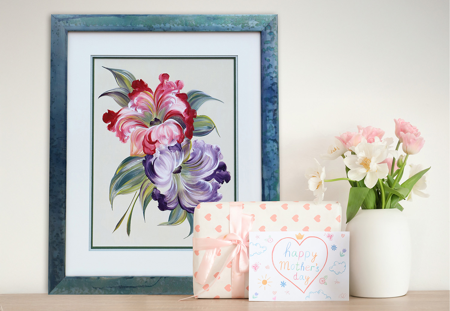 Framed floral artwork with pink and purple flowers displayed beside a Mother’s Day card, wrapped gift, and a vase of fresh flowers on a wooden surface.