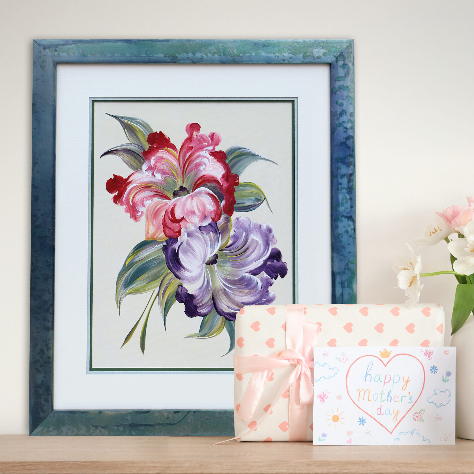 Framed floral artwork with pink and purple flowers displayed beside a Mother’s Day card, wrapped gift, and a vase of fresh flowers on a wooden surface.