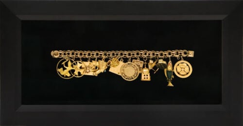 Gold charm bracelet with various intricate pendants displayed in a black shadowbox frame.
