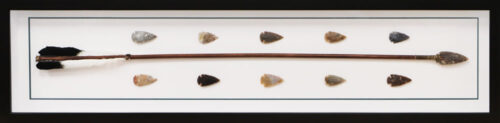 Framed Native American arrow with black and white feathers, surrounded by ten stone arrowheads.
