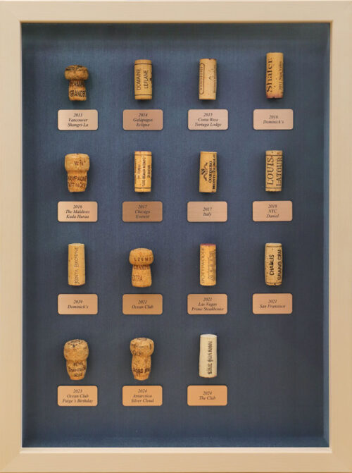 Framed collection of labeled wine corks from global destinations, mounted on navy blue textured matting.