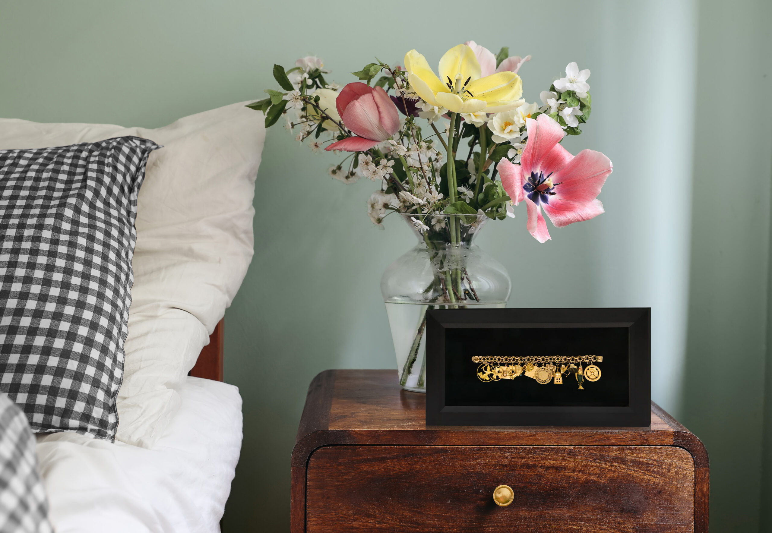 Small shadow box frame displaying a gold charm bracelet mounted against a black velvet backdrop on a wooden nightstand.