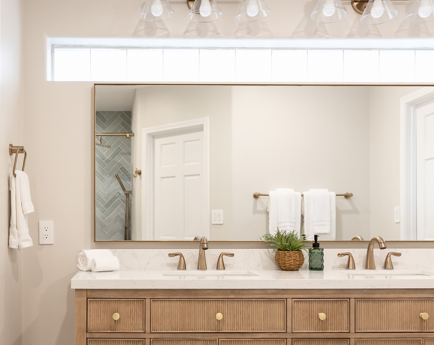 Large horizontal wall mirror with a thin brass metal frame installed over a double-sink bathroom vanity with warm wood cabinets.