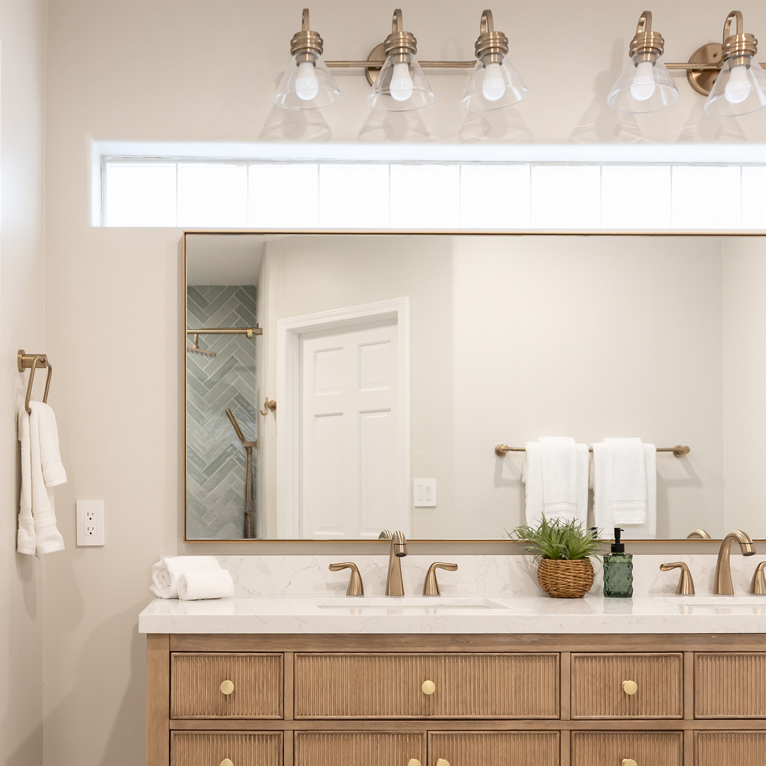 Large horizontal wall mirror with a thin brass metal frame installed over a double-sink bathroom vanity with warm wood cabinets.