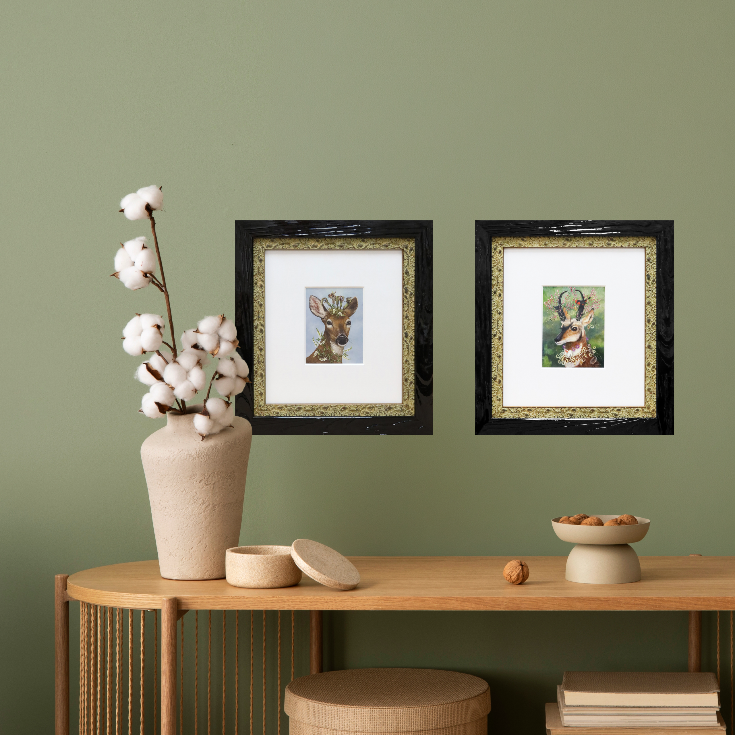 Pair of framed deer art prints with dark wood outer frames and ornate gold inner borders hung on a sage green wall above a wooden console table.