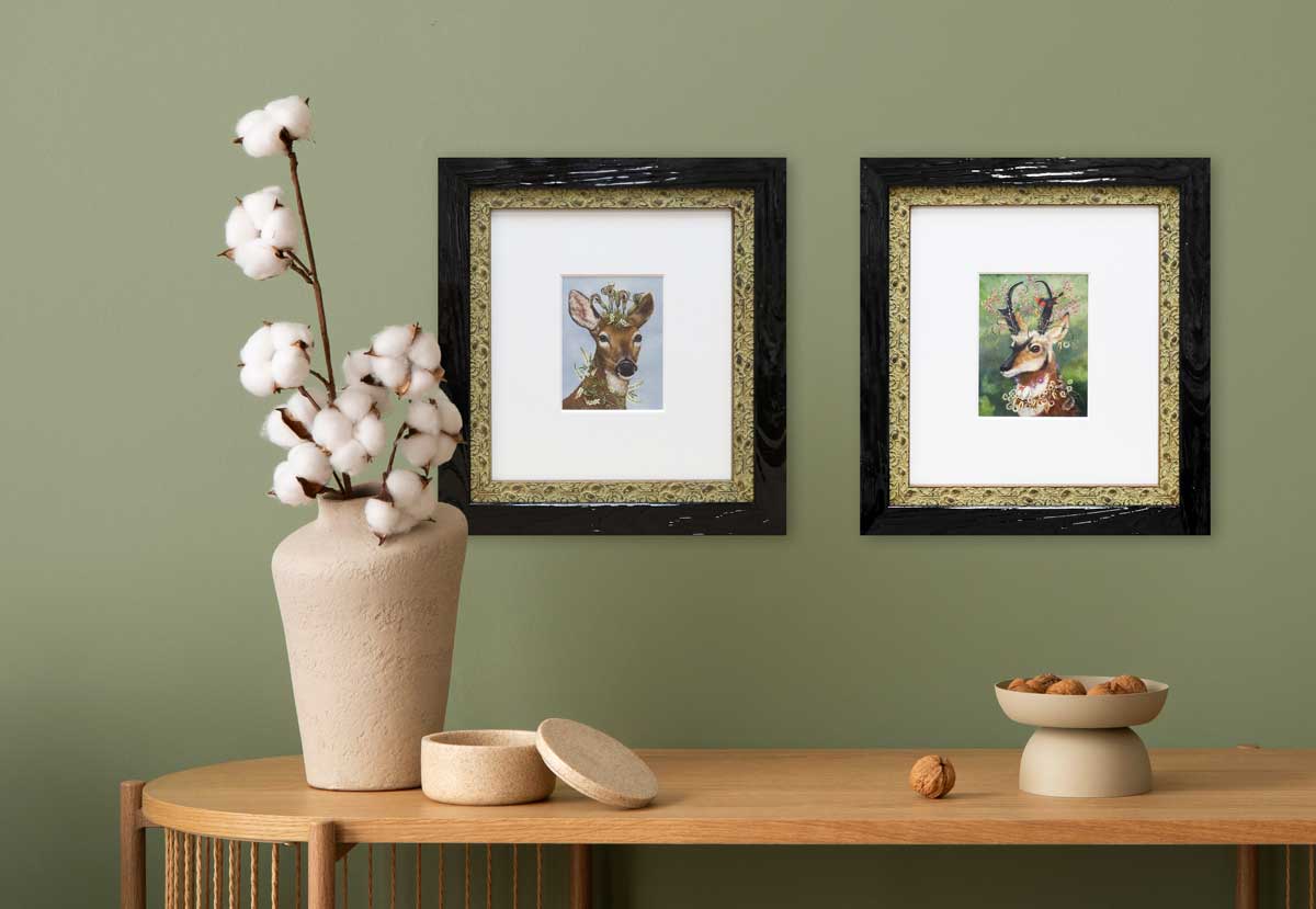 Pair of framed deer art prints with dark wood outer frames and ornate gold inner borders hung on a sage green wall above a wooden console table.