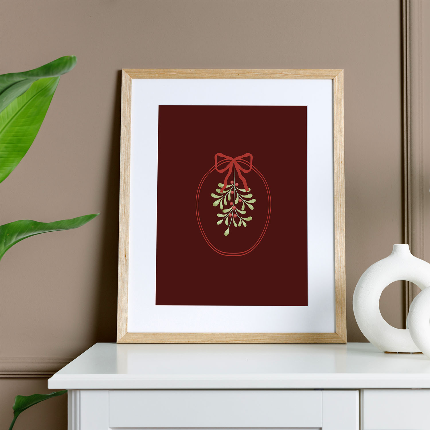 Framed holiday art print of mistletoe with a red ribbon on a deep burgundy background, displayed in a natural light wood ready-made frame with white matting.