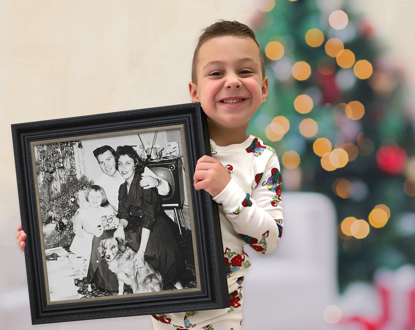 Smiling boy in holiday pajamas holding a framed vintage black-and-white family Christmas photograph in front of a glowing tree.