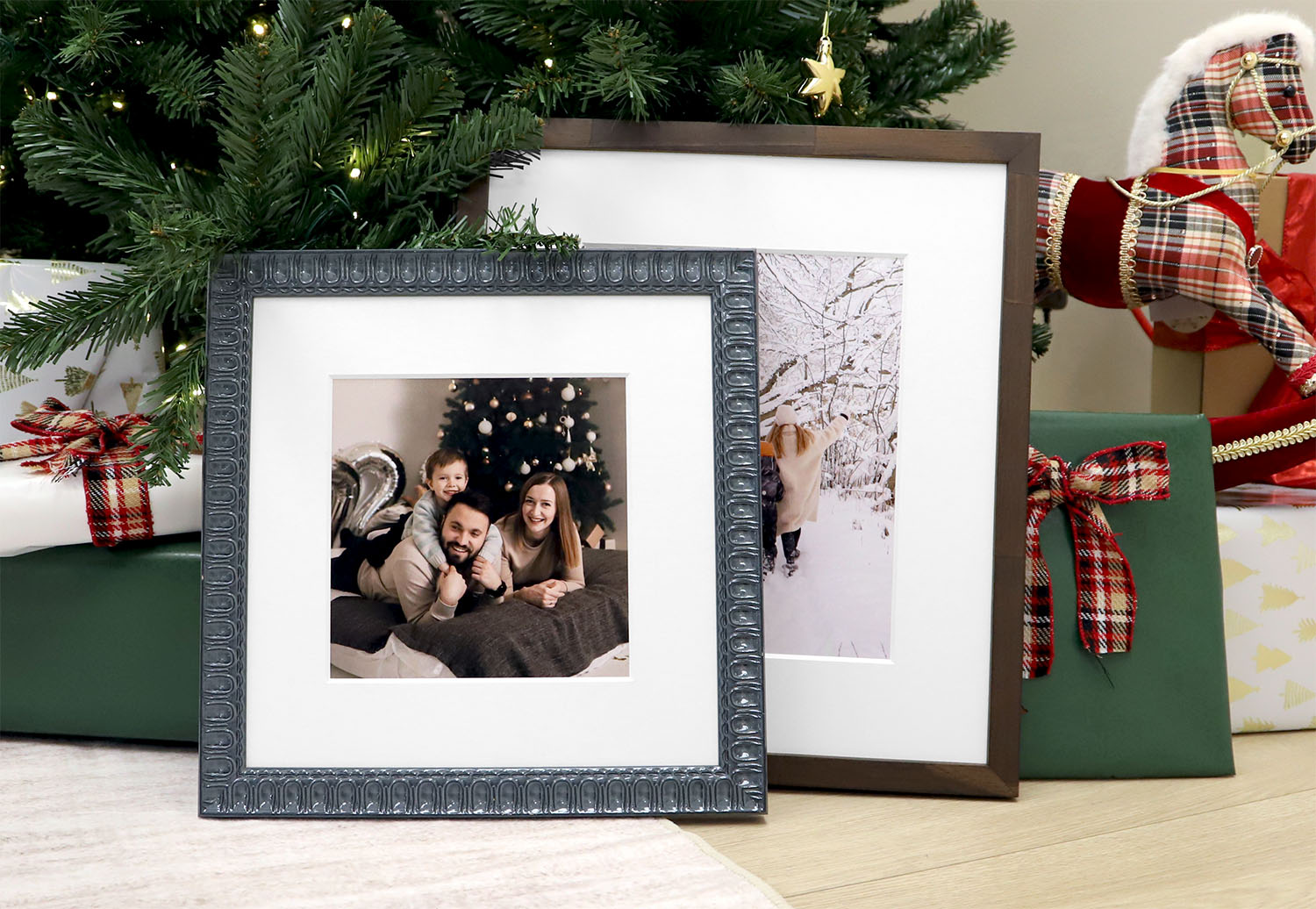 Square framed family portrait in an ornate dark silver frame resting beneath a decorated Christmas tree next to wrapped gifts.