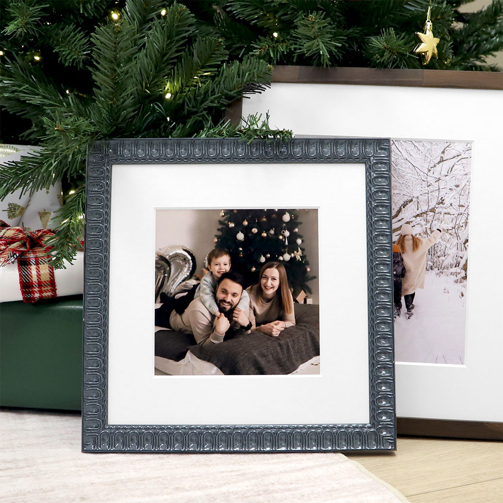 Square framed family portrait in an ornate dark silver frame resting beneath a decorated Christmas tree next to wrapped gifts.